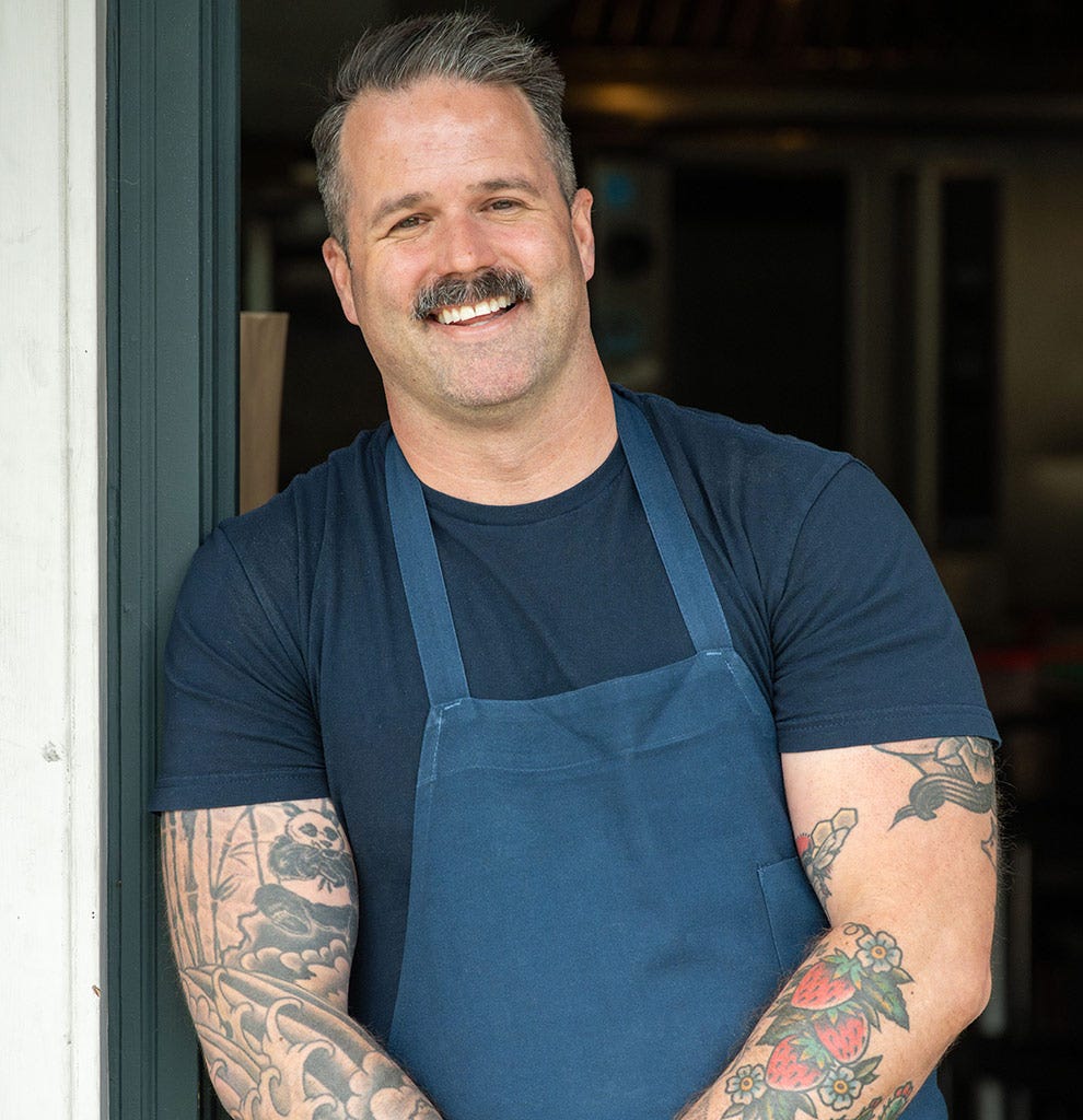 Chef Avery Hardin standing in a kitchen doorway, wearing an apron and smiling. Click to read his interview.