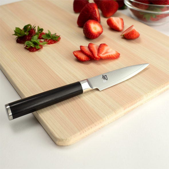 Paring knife beside sliced strawberries on a light wood board.
