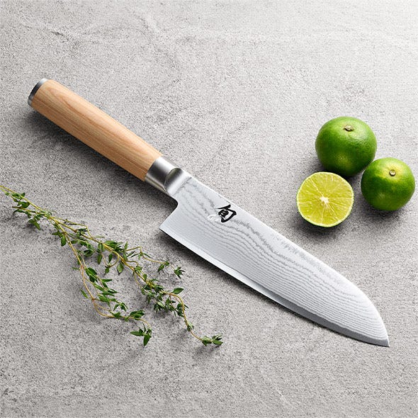 Santoku knife styled with herbs and limes on a stone surface.