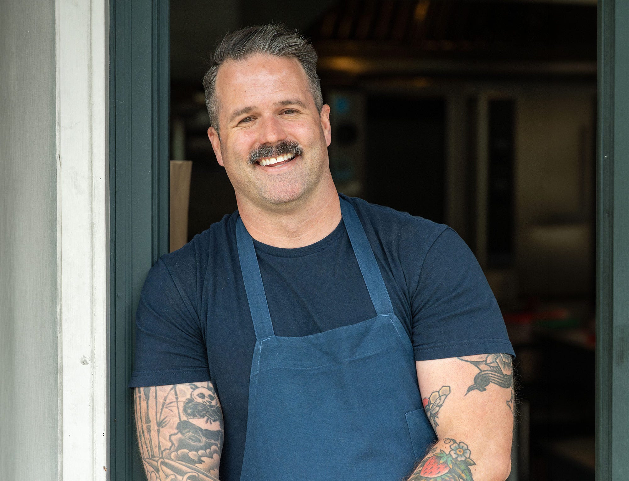 Chef Avery Hardin standing in a kitchen doorway, wearing an apron and smiling.