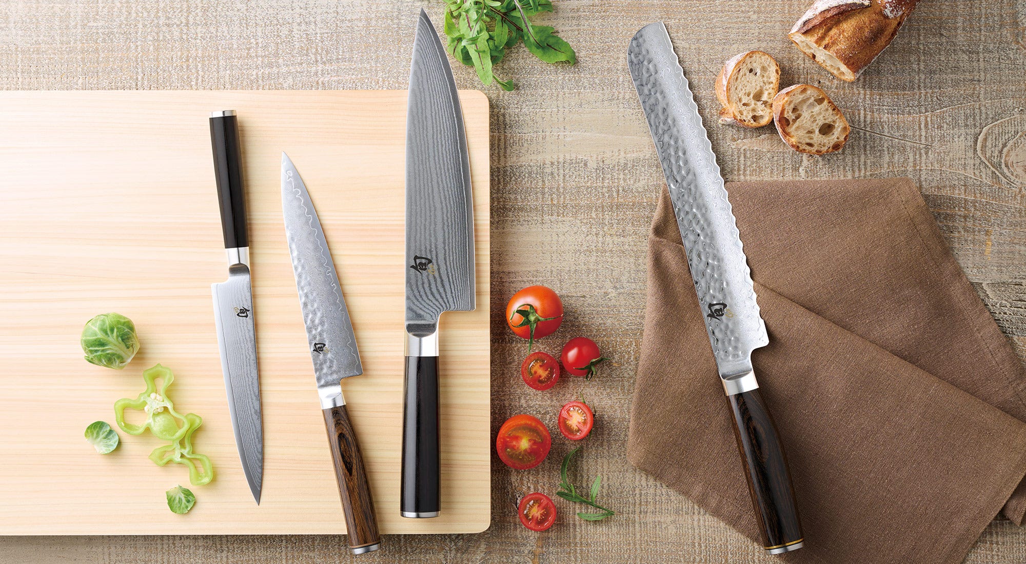 Shun Japanese knives arranged on a Hinoki cutting board with fresh vegetables and bread, demonstrating proper cutting surfaces and knife care.